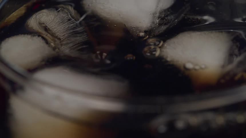 Dark beverage pours into clear glass over floating ice cubes