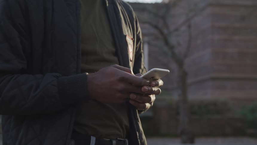 A black man with brown clothing is standing near a brick building typing on his iphone