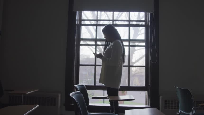 A young woman stands in front of a large window in a classroom while looking down at her phone