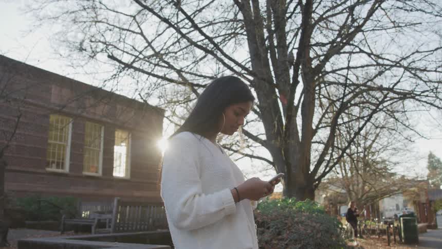 Woman stands outside building viewing mobile phone while others walk through park