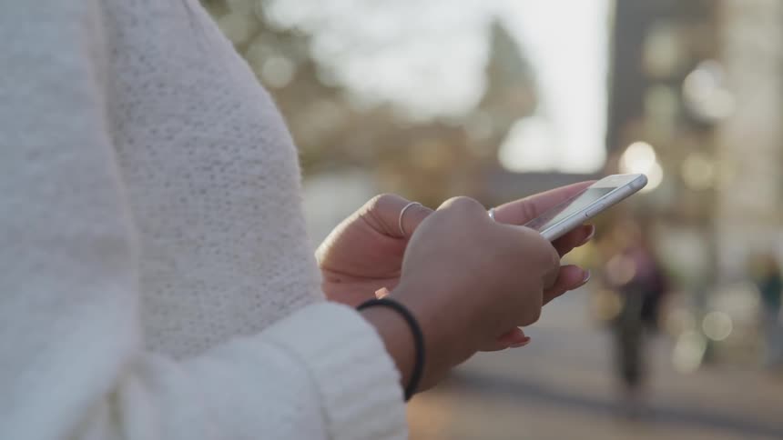 Woman wearing a white sweater types on her phone on the street at sunset