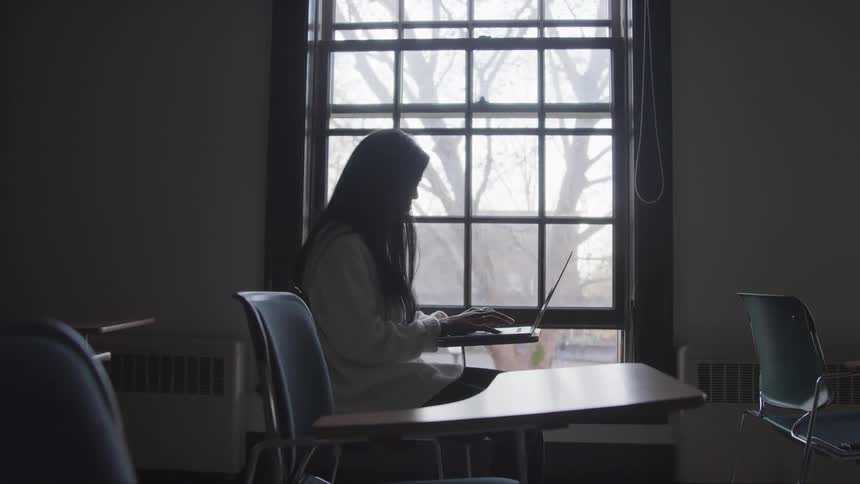 Female student sitting on a desk near a window in a classroom working on a laptop