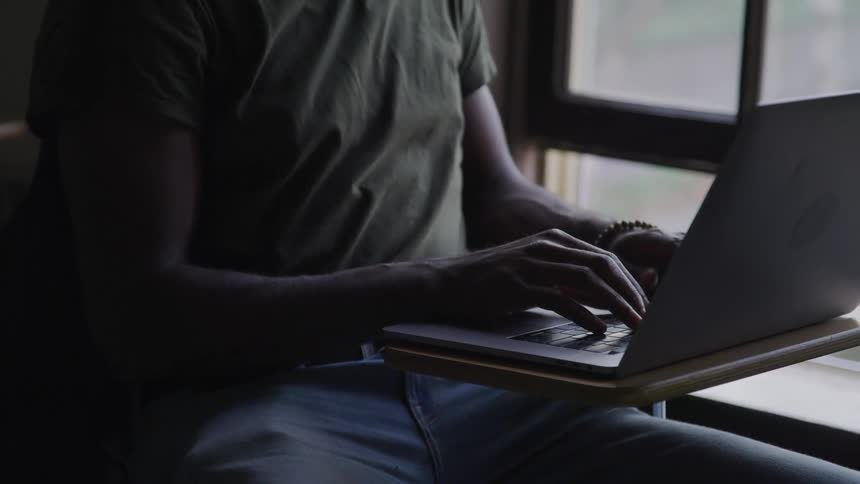 A man sits at a small student desk typing on a laptop