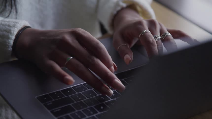 A black woman sits at a desk and types on a laptop using her long fingernails