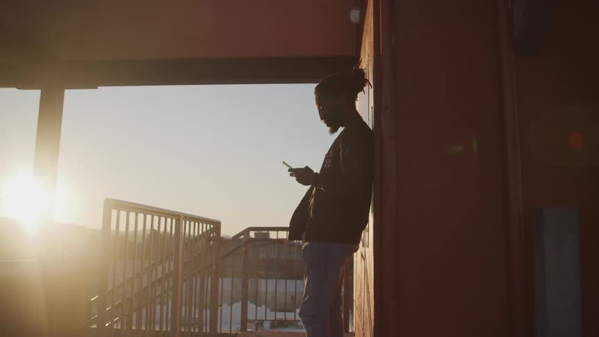 A man standing against a wall holding a smart phone with the sunset in the background