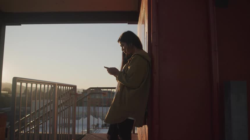 Woman leans against building on upper floor near staircase viewing mobile phone with other buildings in background