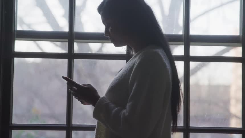 A woman is texting on a cellphone near a window