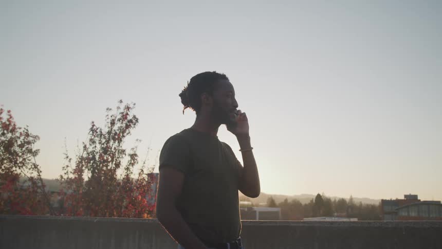 Black man with braid hair a t shirt talking on the smartphone and is silhouetted against the sky on top of a building