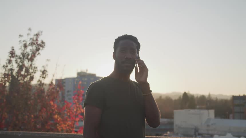 A man stands on a city building balcony holding his phone to his ear and looking concerned