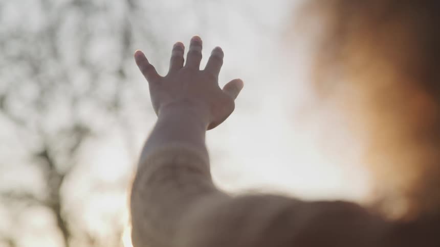 A woman stands outside near barren trees and reaches her arm out to the sun