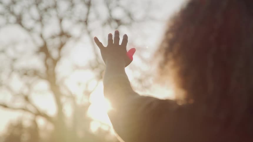 A young woman reaches her hand upwards towards the sun as it shines through the branches of a tree