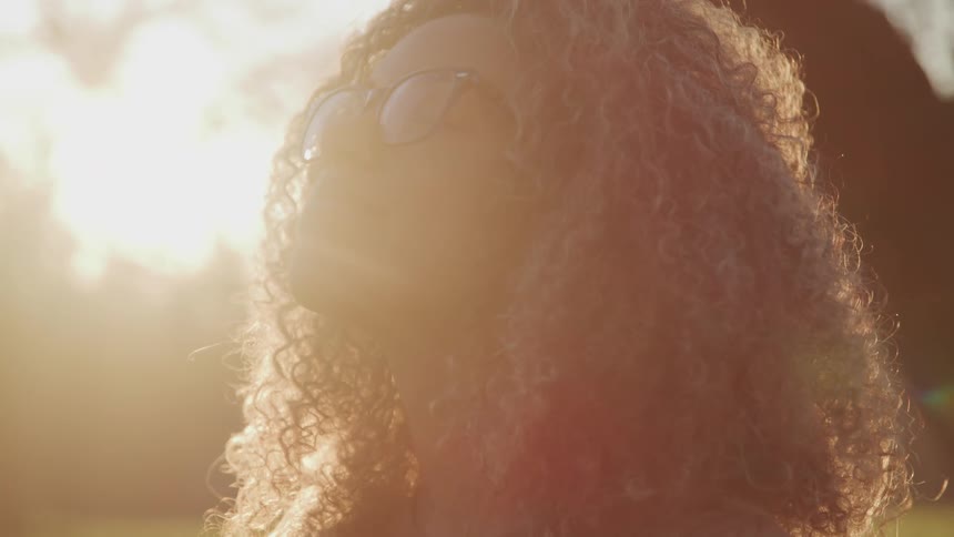 A young woman smiles as she looks upwards and the sky and the sun shines brightly behind her