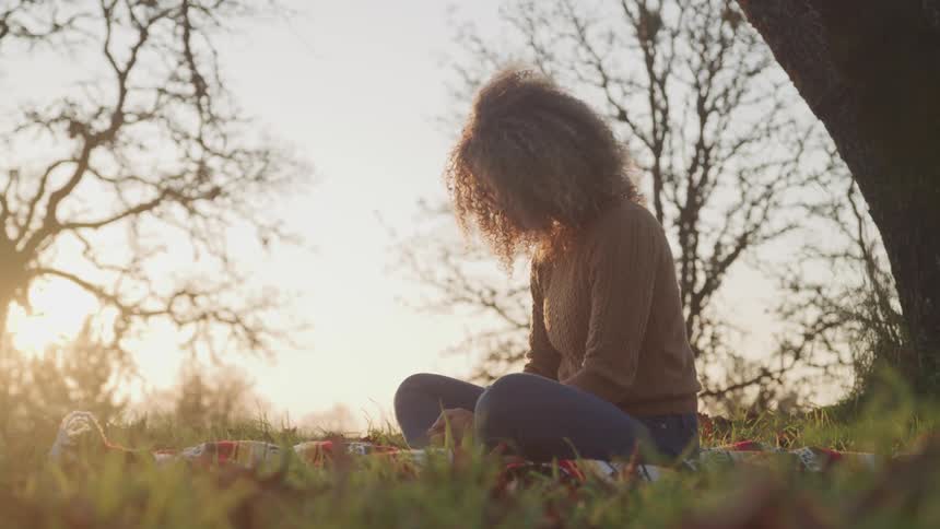 Woman with jeans and a sweater sits cross legged on green lawn at sunset