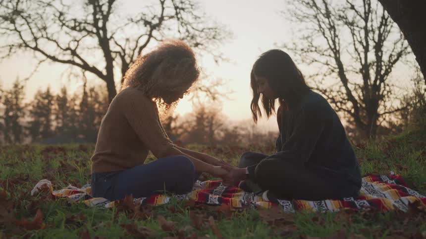 Two women hold hands and pray and they sit on a striped blanket camping in a field near a mountain range