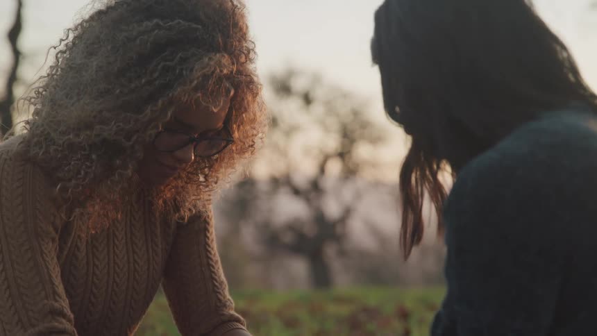 Two women sit facing each other with their heads down in a park