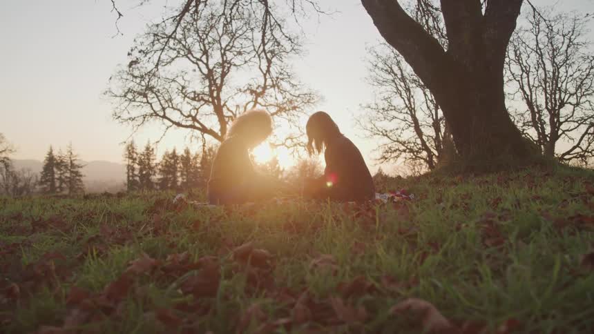 Two women sit on a striped blanket in a field near a mountain and pray with with their heads down