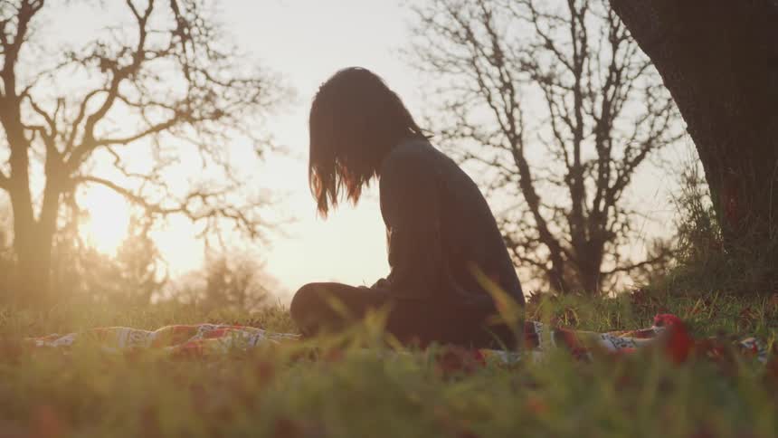 A woman sits on a colorful blanket in the middle of a forest with her head bowed
