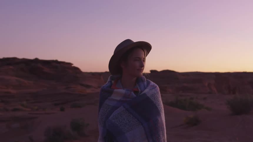Woman wrapped in a blanket and wearing a hat walks on a desert looking pensively at the horizon at sunset