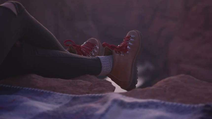 A woman sits on a blanket on the ground with her feet hanging over the edge of canyon in the evening