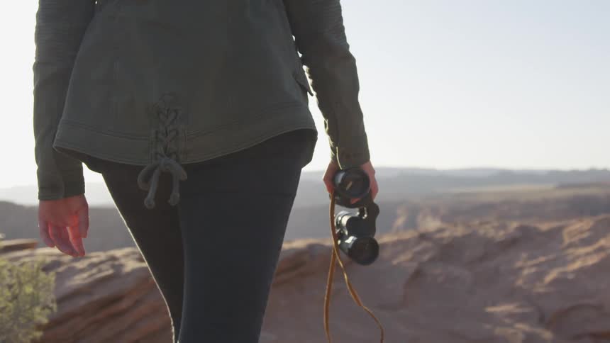 A woman walks across a tall dry terrain holding binoculars