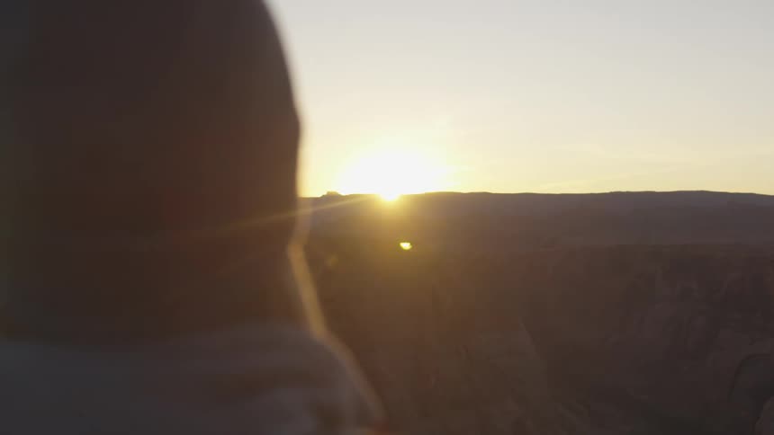 A woman with a hat standing by a canyon with the sky and the sun in the background