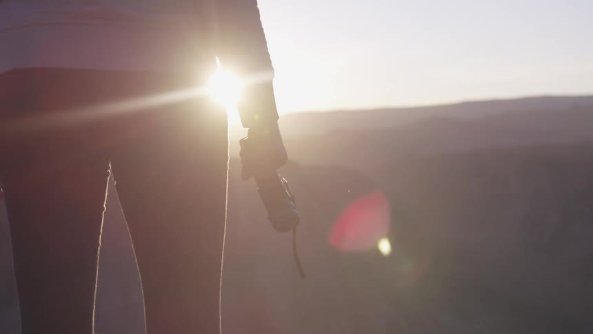 A person standing with a camera in their hand with a canyon the sky and the sun in the background