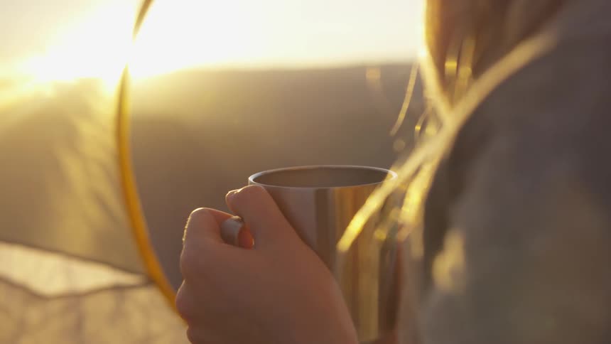 Woman camping on a tent looks at the sunset while drinking a beverage