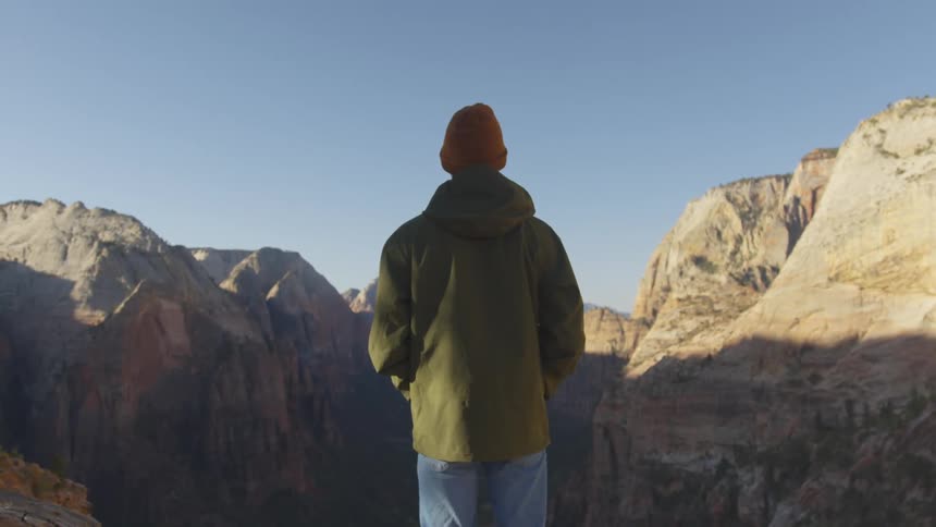 Person wearing knit hat and coat stands looking at shadowy canyon and surrounding cliffs under blue sky