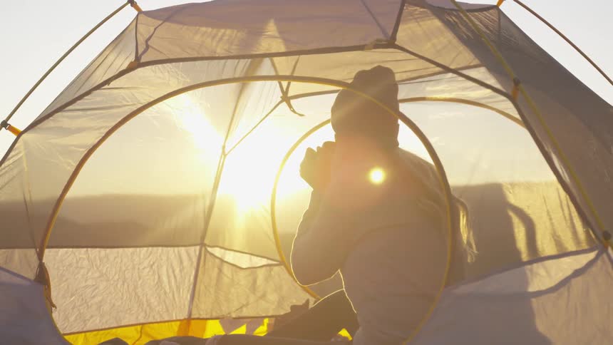A woman sits in a tent and drinks from a mug