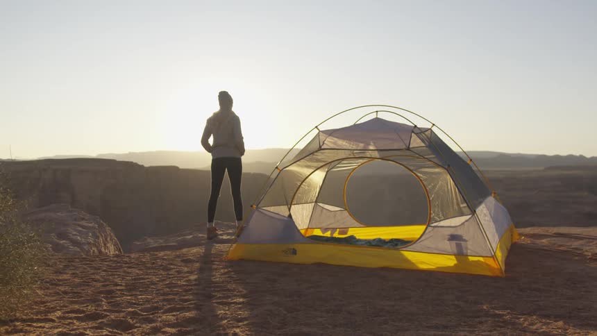 A woman stands outdoors next to her camping tent as she enjoys the landscape around her