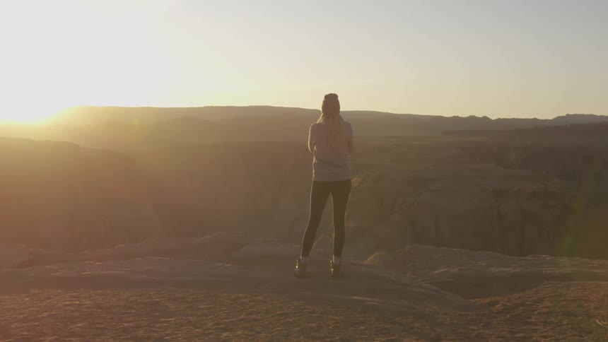 A woman with a hat standing by a canyon with the sky and sun in the background