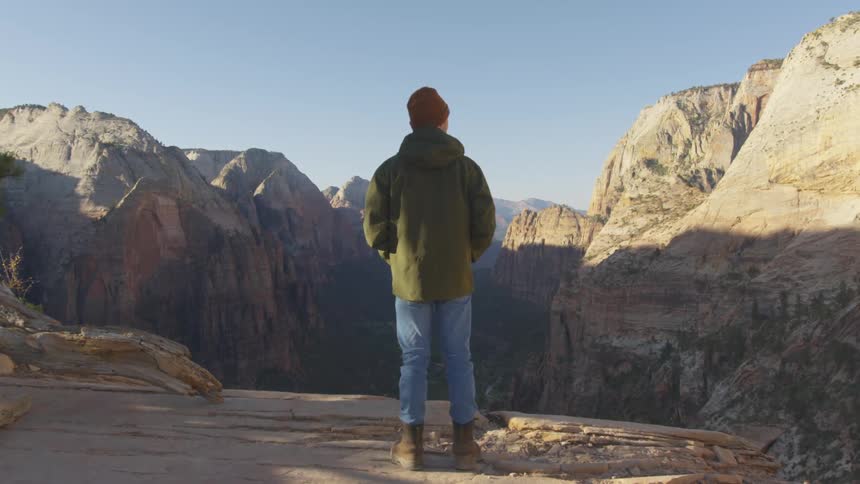 A man with a hat a coat and boots standing by a cliff with a canyon in the background