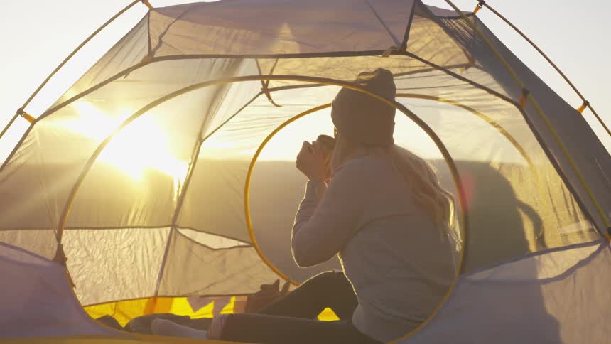 Young woman wearing a sweater drinks hot beverage on a tent in the mountains