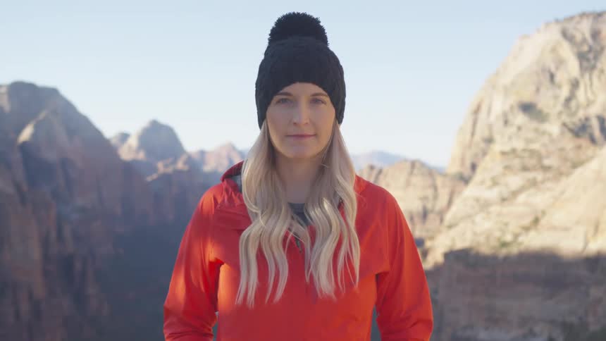 A woman standing high in the mountains on a sunny day slowly raises her head to look into the camera