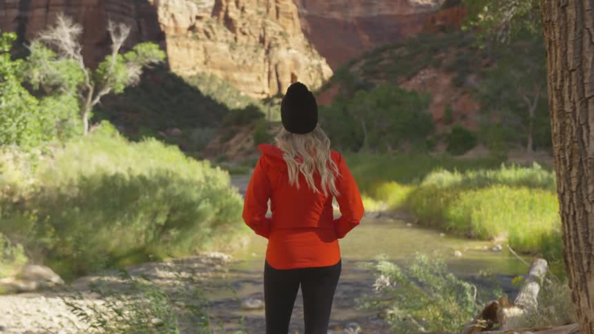 A woman with a hat and jacket standing with a cliff and trees in the background