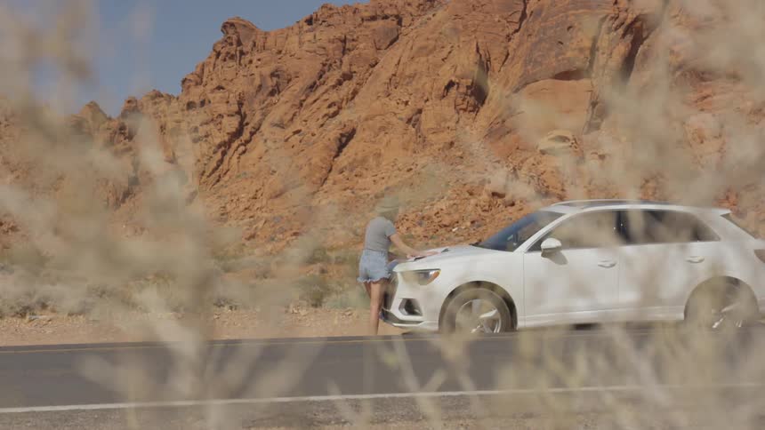 A woman with a hat standing in front of a car by a road with a cliff and the sky in the background
