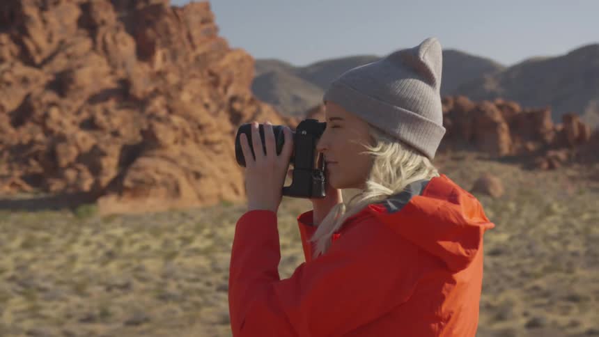 A woman stands in desert terrain and holds a camera up to her face