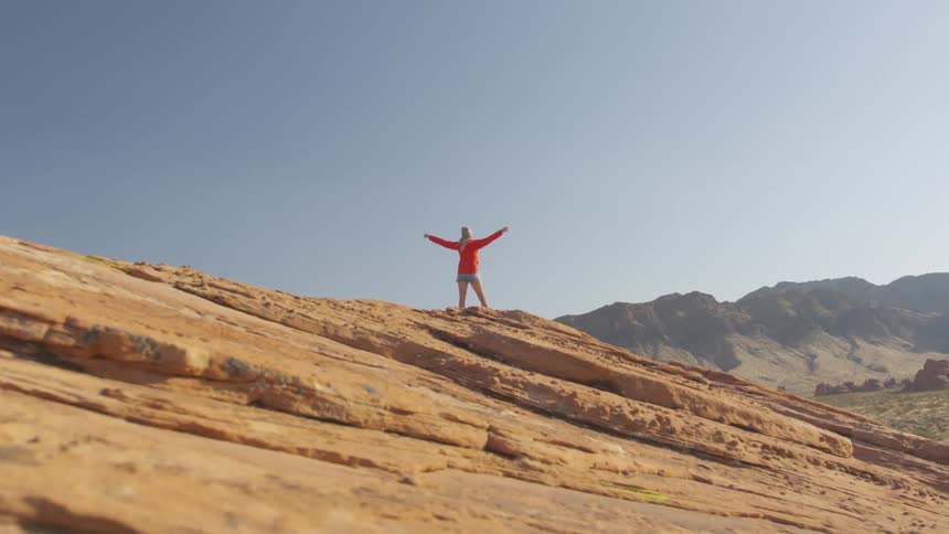 A woman with a hat standing on a cliff with a mountain range and the sky in the background