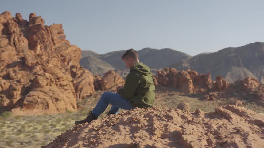 Man wearing a hooded jacket sits on the rocks as mountains sit in the distance