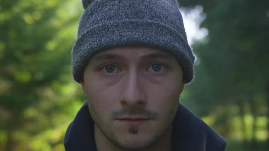 Man with a beard and a grey beanie raises his head and looks at the camera in a forest