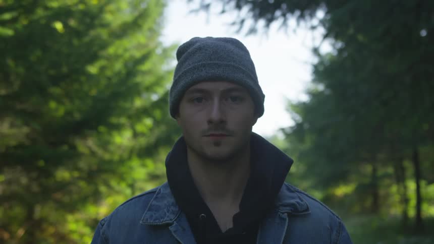 A man with a hat and jacket standing and staring with trees and the sky in the background