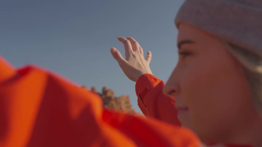 Young blonde woman wearing a beanie and a red jacket raises her hands in the wind on the desert