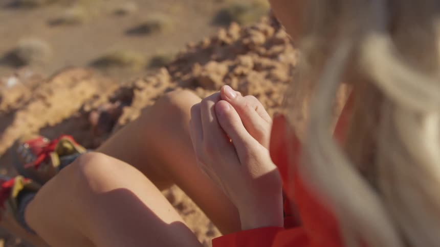 Blonde woman sits with clasped hands on sand as wind ruffles her hair