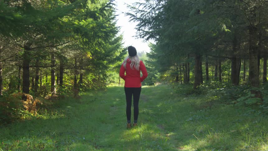 Woman with long blonde hair a black beanie and a red coat stands on a trail through the forest