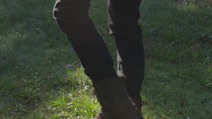 A thin person wearing tall boots walks over a grassy field