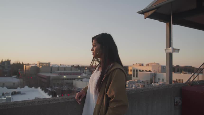 A young woman stands on the rooftop of a building while looking out over the city below lost in thought