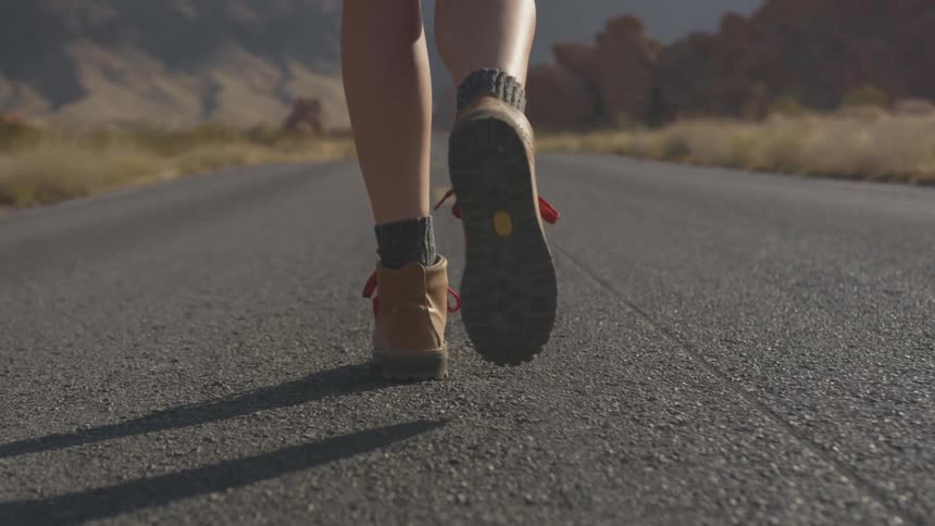 A white person in hiking boots is waking slowly down a empty road in the desert