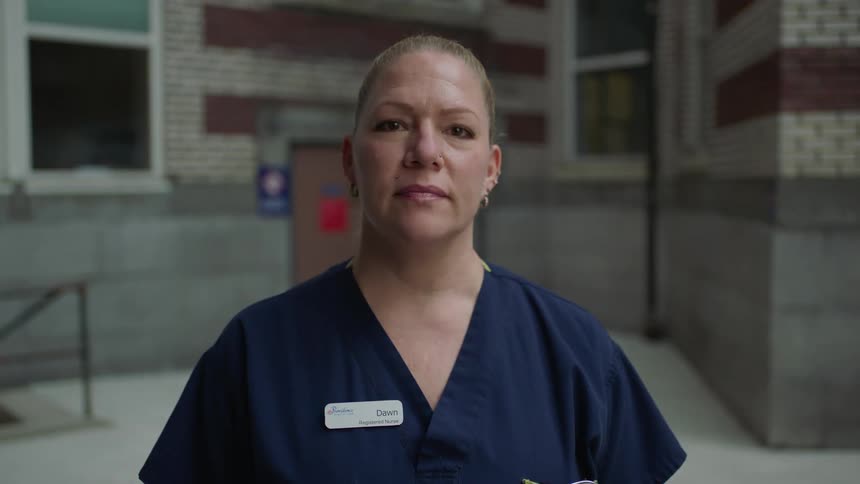 A white woman wearing scrubs stands outside a building