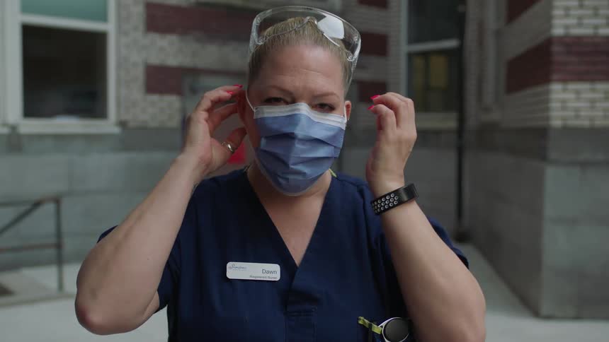 A white female medical worker stands outside a building and puts a face mask on