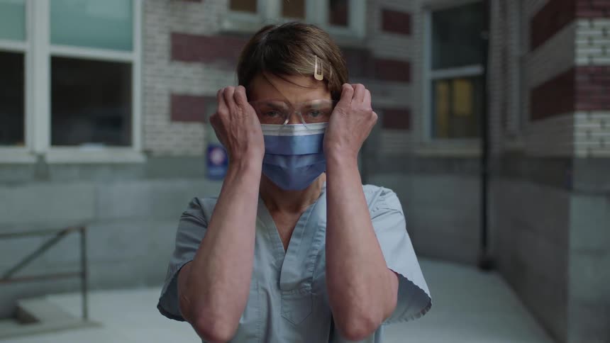 A woman with blue scrubs and a disposable mask putting on glasses